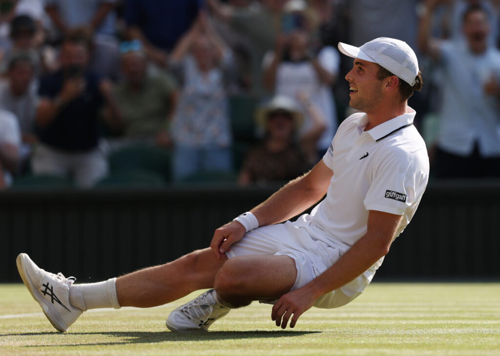 Tennis player in white kit sliding on a grass court, smiling, wearing a white cap, with spectators blurred in the background