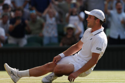 Tennis player in white kit sliding on a grass court, smiling, wearing a white cap, with spectators blurred in the background