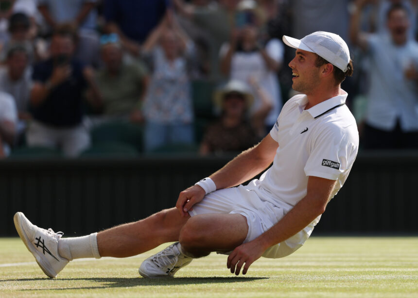 Tennis player in white kit sliding on a grass court, smiling, wearing a white cap, with spectators blurred in the background