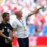 Soccer referees on the field; one in black holding a ball, the other in a white shirt pointing toward the stands.