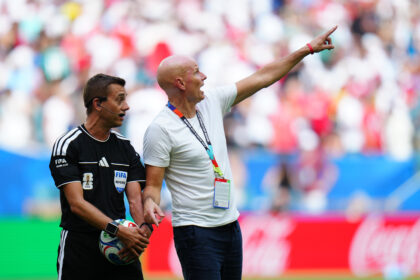 Soccer referees on the field; one in black holding a ball, the other in a white shirt pointing toward the stands.