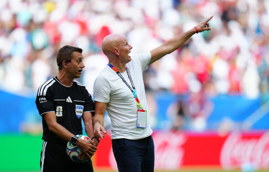 Soccer referees on the field; one in black holding a ball, the other in a white shirt pointing toward the stands.