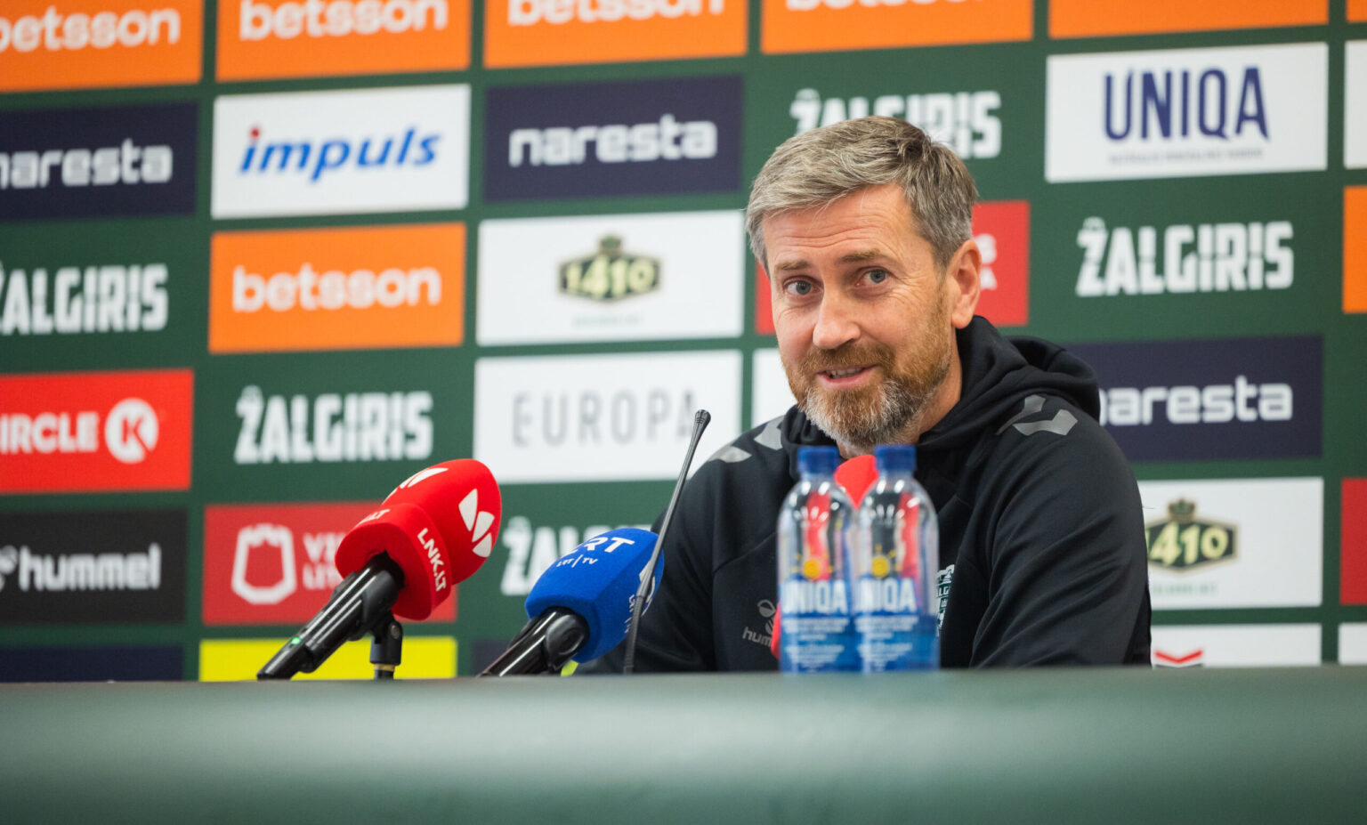 Man with gray hair and beard sits at a press conference, microphones in the foreground and sponsor logos on a colorful wall behind him.