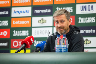 Man with gray hair and beard sits at a press conference, microphones in the foreground and sponsor logos on a colorful wall behind him.