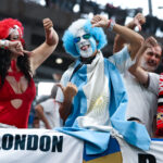 Group of sports fans in colorful costumes, raising their arms and cheering at a stadium with a London banner visible, blue and white outfits prominent.