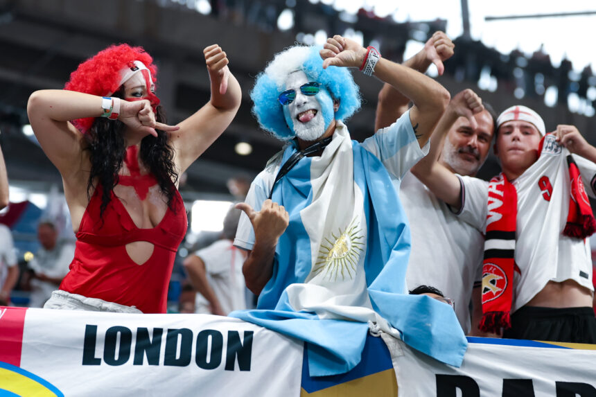 Group of sports fans in colorful costumes, raising their arms and cheering at a stadium with a London banner visible, blue and white outfits prominent.