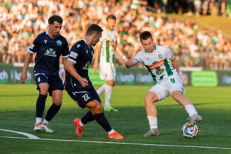Players contesting a soccer ball on a green pitch as two in navy kits close in, with a white-green team member nearby and a packed stadium in the background.