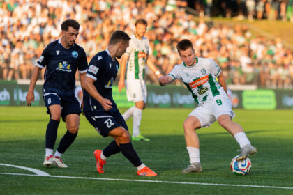 Players contesting a soccer ball on a green pitch as two in navy kits close in, with a white-green team member nearby and a packed stadium in the background.