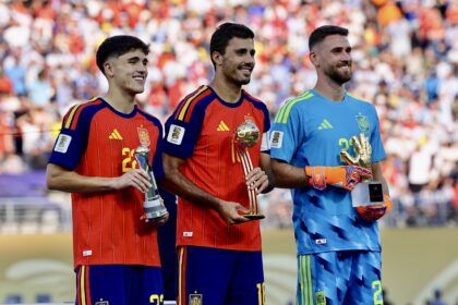 Three male soccer players in red and blue Spain uniforms posing with trophies on a podium in front of a cheering crowd, smiling.