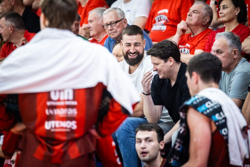 Smiling man with a beard in a white shirt among cheering spectators in red jerseys at a sports event.