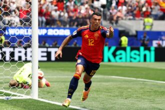 Soccer player in red jersey (Spain) scoring near the goal as the ball crosses the line; goalkeeper on the ground inside the net with crowd in the background.