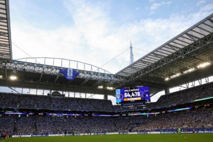 Large soccer stadium with a metal roof, stadium lights, and a big video scoreboard for FIFA World Cup 2026, filled with fans under a blue sky