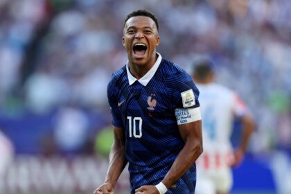 French footballer celebrating a goal on the pitch, fists clenched and mouth open in triumph with a blurred crowd behind him.