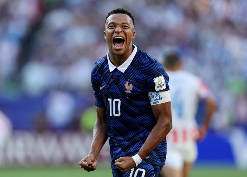 French footballer celebrating a goal on the pitch, fists clenched and mouth open in triumph with a blurred crowd behind him.