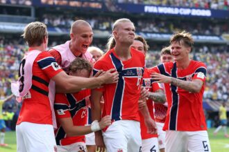 Norwegian national team players celebrate a goal on the field, arms around each other, in red uniforms.