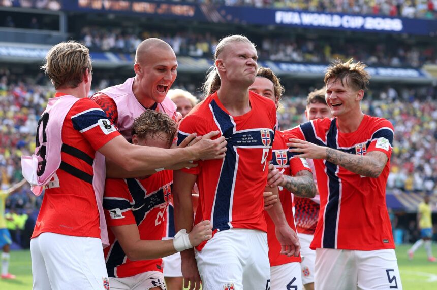 Norwegian national team players celebrate a goal on the field, arms around each other, in red uniforms.