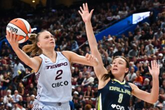 Two female basketball players reach for the ball during a game in a crowded arena, mid-action FIBA-style play.