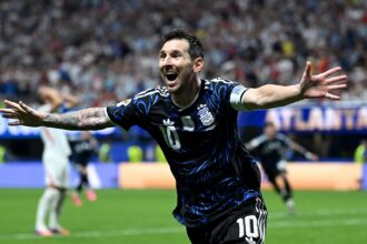 Soccer player in a blue-black jersey with number 10 celebrates with arms outstretched on the pitch amid a cheering crowd.