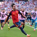 Soccer player in a red and yellow Spain kit sprinting on a green pitch with cheering fans in the stands behind him.