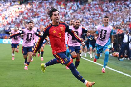 Soccer player in a red and yellow Spain kit sprinting on a green pitch with cheering fans in the stands behind him.