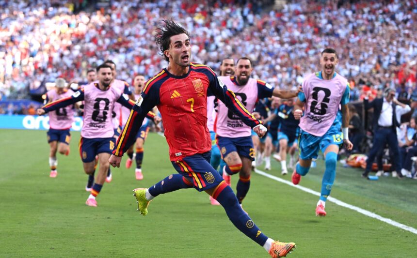 Soccer player in a red and yellow Spain kit sprinting on a green pitch with cheering fans in the stands behind him.