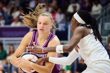 Two female basketball players clash for the ball during a game, one in maroon holding a ball while the other in white defends closely.