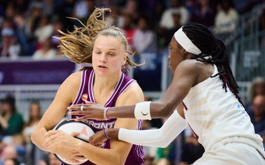 Two female basketball players clash for the ball during a game, one in maroon holding a ball while the other in white defends closely.