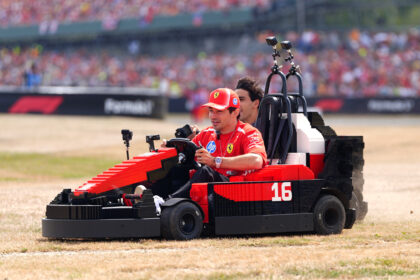 Man in a red Ferrari uniform driving a red LEGO-like race car on a dirt track with a blurred crowd in the stands behind him.
