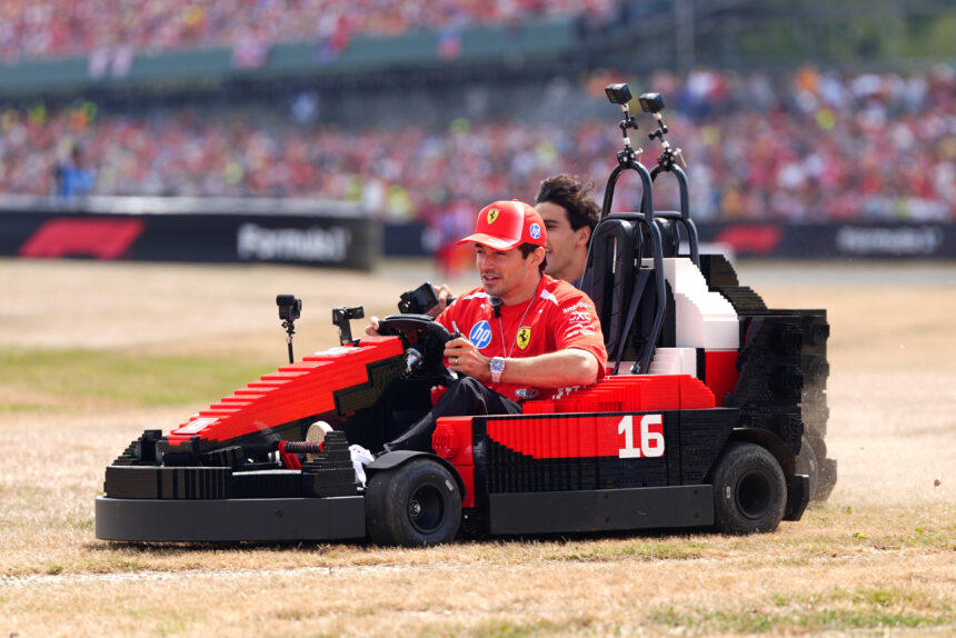 Man in a red Ferrari uniform driving a red LEGO-like race car on a dirt track with a blurred crowd in the stands behind him.