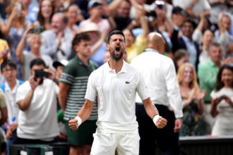 A man in a white polo shouts with clenched fists as a crowd applauds behind him at a sporting event