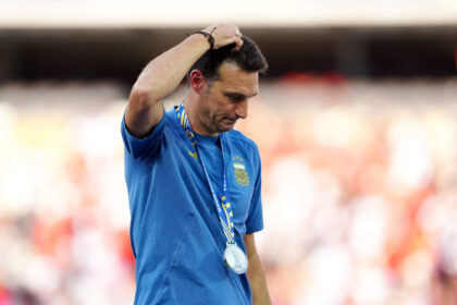 Man in a blue shirt with a medal around his neck, hand resting on his head, standing in front of a blurred crowd at a stadium.