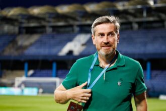 Man in a green polo with an 'All Access' badge on a lanyard at a soccer stadium, posing on the field.