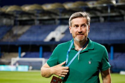 Man in a green polo with an 'All Access' badge on a lanyard at a soccer stadium, posing on the field.
