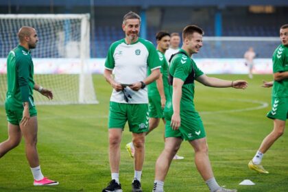 Soccer team in green uniforms on a training field, coach with clipboard smiling among players near a goal.