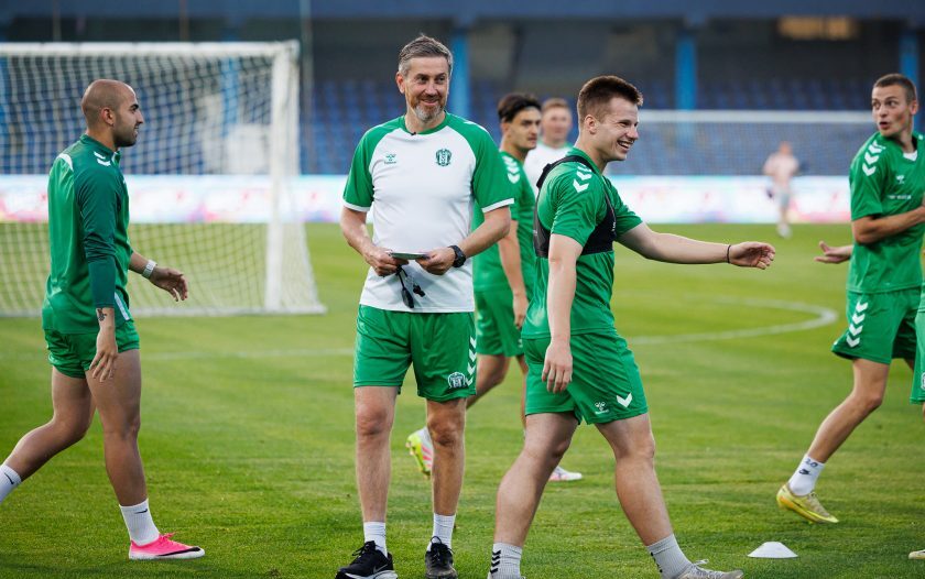 Soccer team in green uniforms on a training field, coach with clipboard smiling among players near a goal.