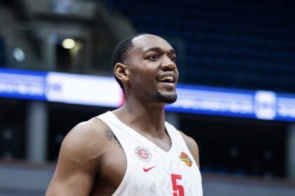 Basketball player in a white jersey with the number 5, smiling on an indoor court with blue signage in the background.
