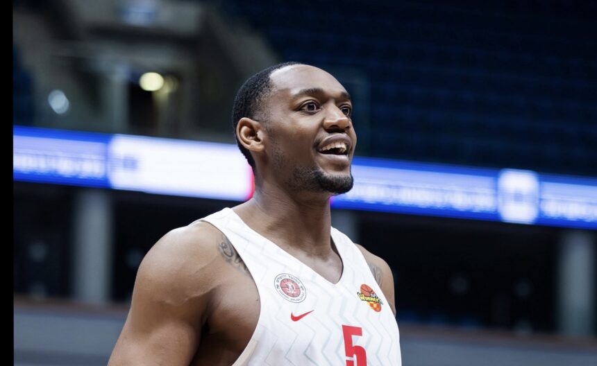 Basketball player in a white jersey with the number 5, smiling on an indoor court with blue signage in the background.