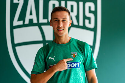 Male soccer player in a green jersey points to his team crest on the chest, with a large green logo backdrop behind him.