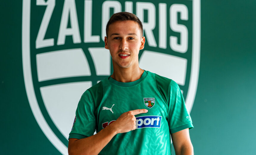 Male soccer player in a green jersey points to his team crest on the chest, with a large green logo backdrop behind him.