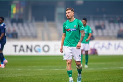 Soccer player in a green jersey walks on a grassy field during a match, spectators blurred in stands behind him.