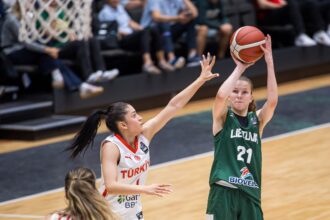 Lithuanian player in green (#21) shoots the ball while a defender in white reaches up to block, in a gym with blurred spectators behind them.