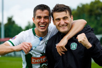 Two smiling soccer players with arms around each other celebrate on a grassy field, one in a white jersey and the other in a black jacket.