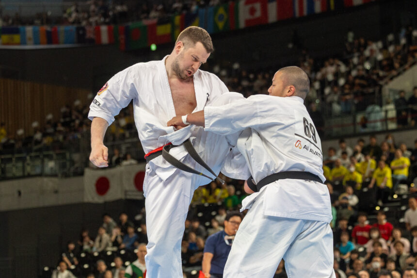 Two men in white martial arts uniforms sparring in a packed arena, with spectators in the stands behind them.