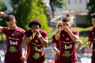 Four soccer players in maroon jerseys celebrate on a sunny field, raising fists and clapping.
