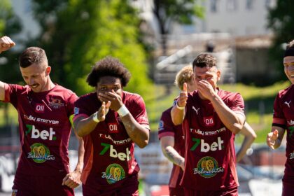 Four soccer players in maroon jerseys celebrate on a sunny field, raising fists and clapping.