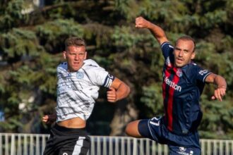 Two soccer players compete for the ball in mid-air; a white kit player on the left and a navy kit with a red stripe on the right.