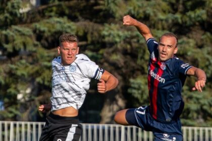 Two soccer players compete for the ball in mid-air; a white kit player on the left and a navy kit with a red stripe on the right.