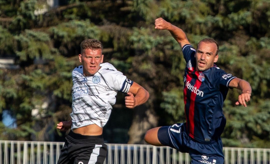Two soccer players compete for the ball in mid-air; a white kit player on the left and a navy kit with a red stripe on the right.