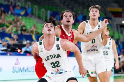 Lithuania national basketball players in white jerseys reach upward during a game as green arena seats and crowd loom in the background, #32 in front.