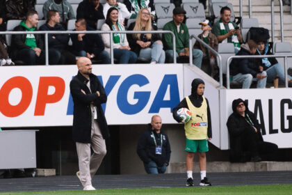 Coach with arms crossed on the sideline as a youth player in a yellow bib holds a soccer ball nearby; crowd in the stands behind them.